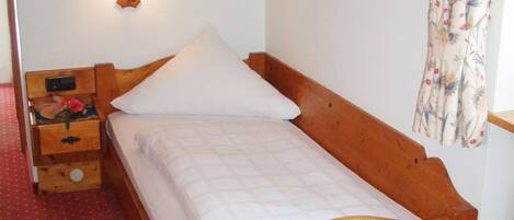 Una cama individual con cabecera de madera, una mesita de noche con lámpara y una cortina estampada con motivos florales.