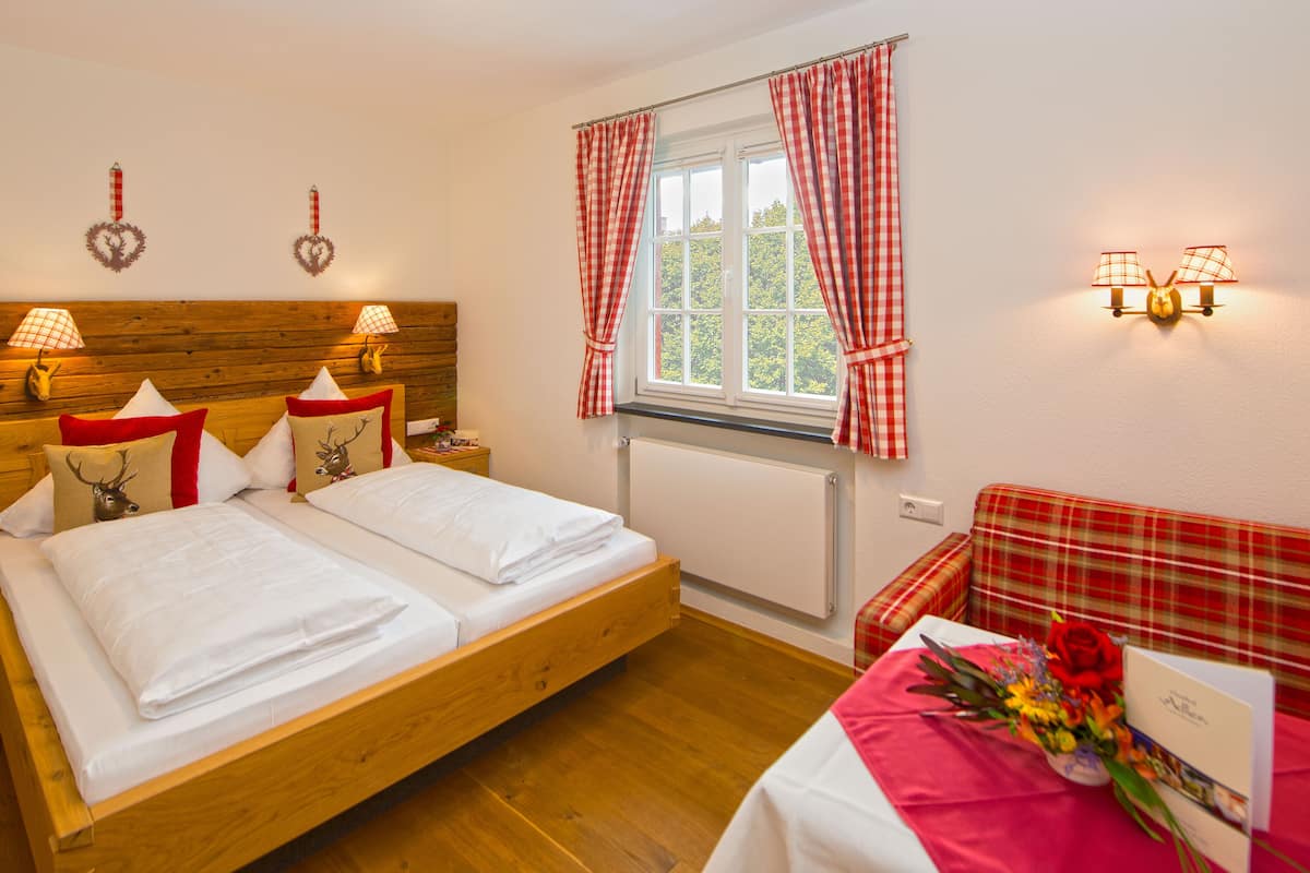 Een tweepersoonsbed met witte lakens, een houten hoofdbord, twee bedlampen, een raam met rood geruit gordijnen, een geruit bankje en een tafel met een rode tafelkleed en bloemen.