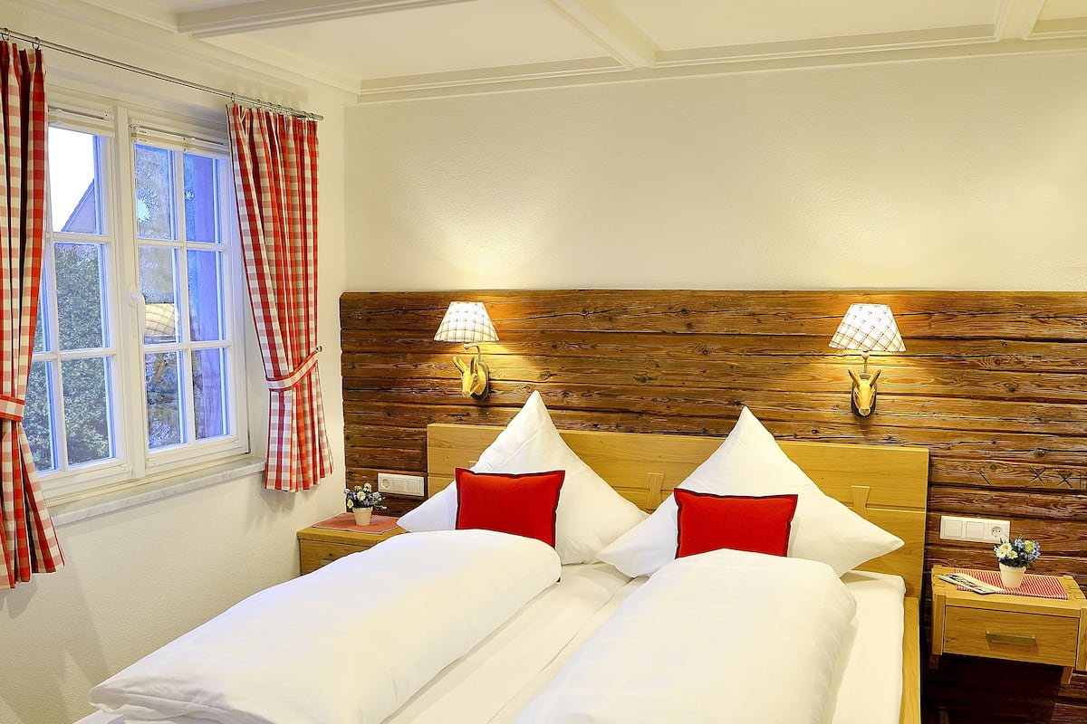Een tweepersoonsbed met wit en rode beddengoed, twee bedlampen, een houten hoofdbord en een raam met rood-witte gordijnen.