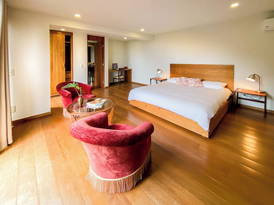 Un dormitorio espacioso con una cama grande, suelo de madera, un sillón rojo, una mesita con libros y un escritorio con una silla.