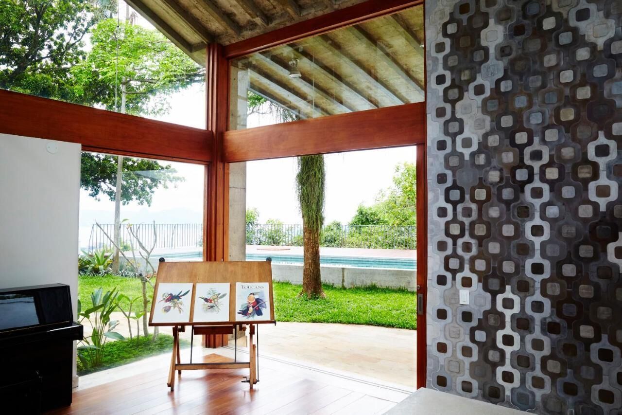 A room with a wooden floor, a painting on an easel, a piano, and a view of the outdoors through large windows.