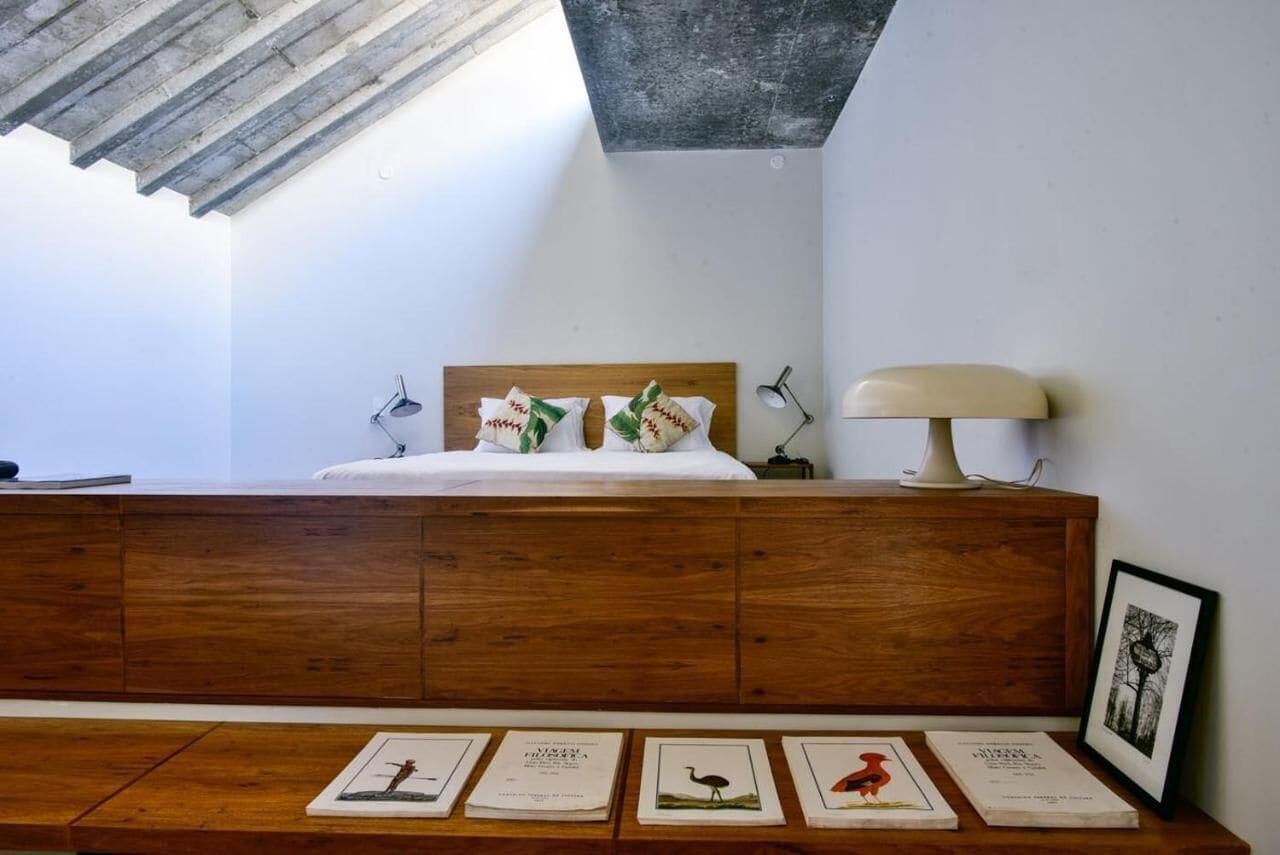 Un dormitorio con cabecera de madera, una mesita de noche con lámpara y un tragaluz sobre la cama.