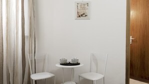 A small room with a white door, a white wall, a small table with two white chairs, and a framed picture above the table. - A room close to Colosseum (Roma)