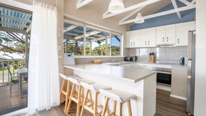 Een moderne keuken met witte kasten, een groot eiland met barkrukken, een inbouwoven en uitzicht op de omgeving door grote ramen.