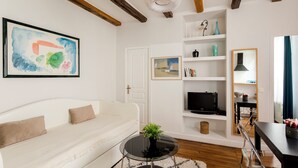 A modern living room with a white sofa, a glass coffee table with a plant, a TV on a shelf, and a wooden ceiling. - Sévigné le Marais Place des Vosges (Paris)