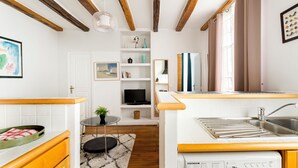 A compact kitchen with wooden beams, a built-in oven, a sink, and a small dining area with a table and chair. - Sévigné le Marais Place des Vosges (Paris)
