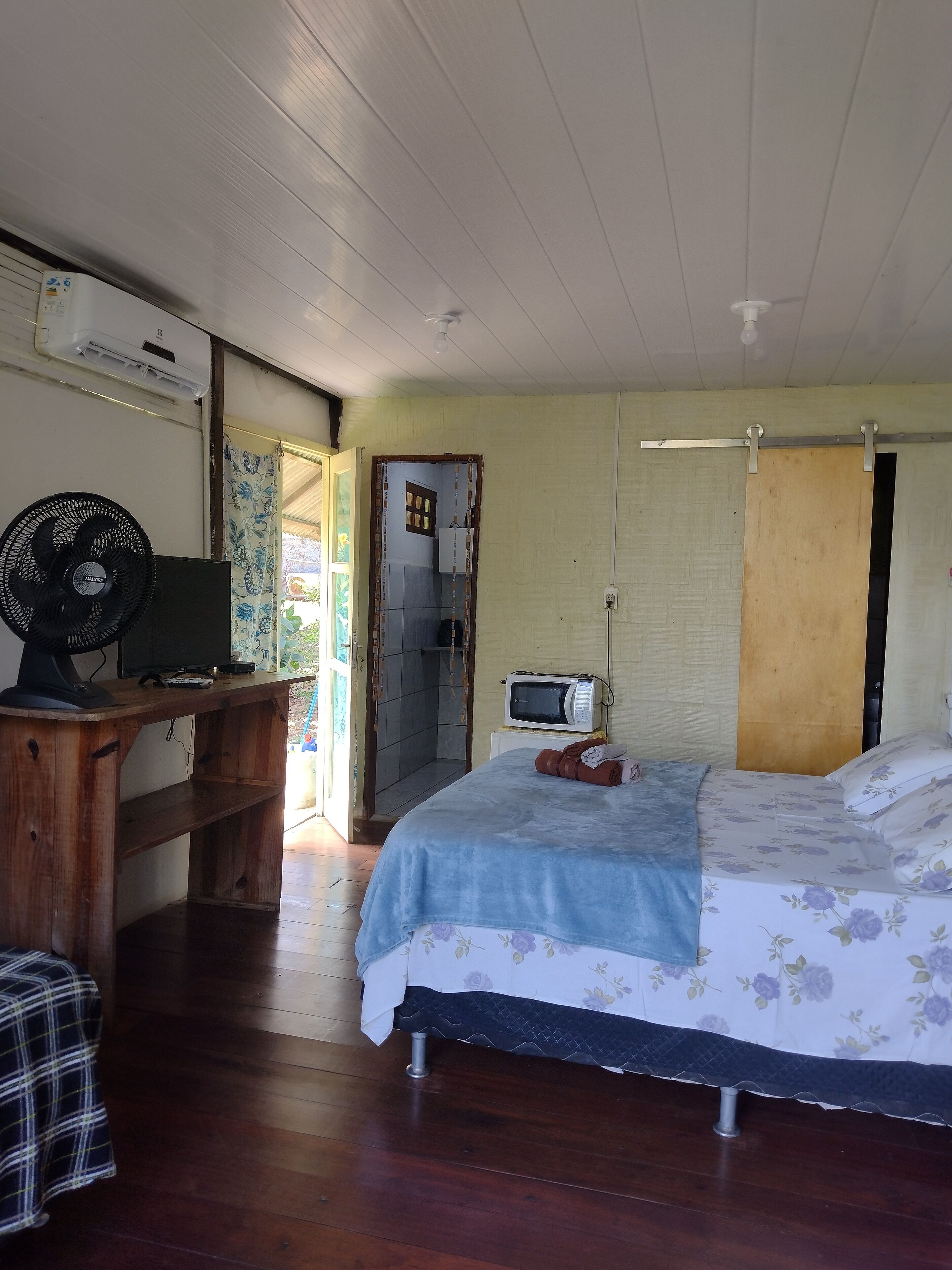 A room with a bed, a desk with a computer and fan, a microwave, and a shower area.