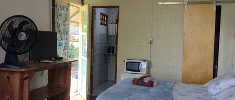 A room with a bed, a desk with a computer and fan, a microwave, and a shower area.