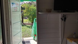 A room with a television on a stand, a door leading outside, and a small shelf with a bottle. - Palhoça da Colina (Fernando de Noronha)