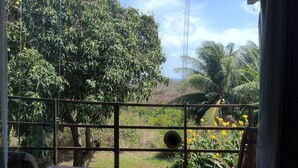 A wooden deck with two lounge chairs, a metal railing, and a view of a lush garden with trees and yellow flowers. - Palhoça da Colina (Fernando de Noronha)