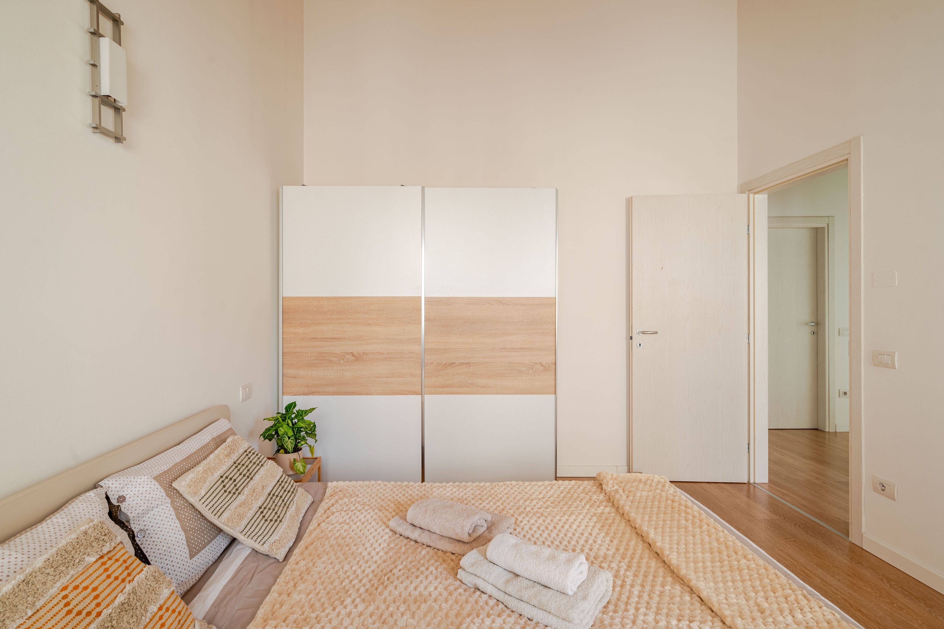 Un dormitori amb un llit, una taula de nit amb una planta, un armari i una porta que condueix a una altra habitació.