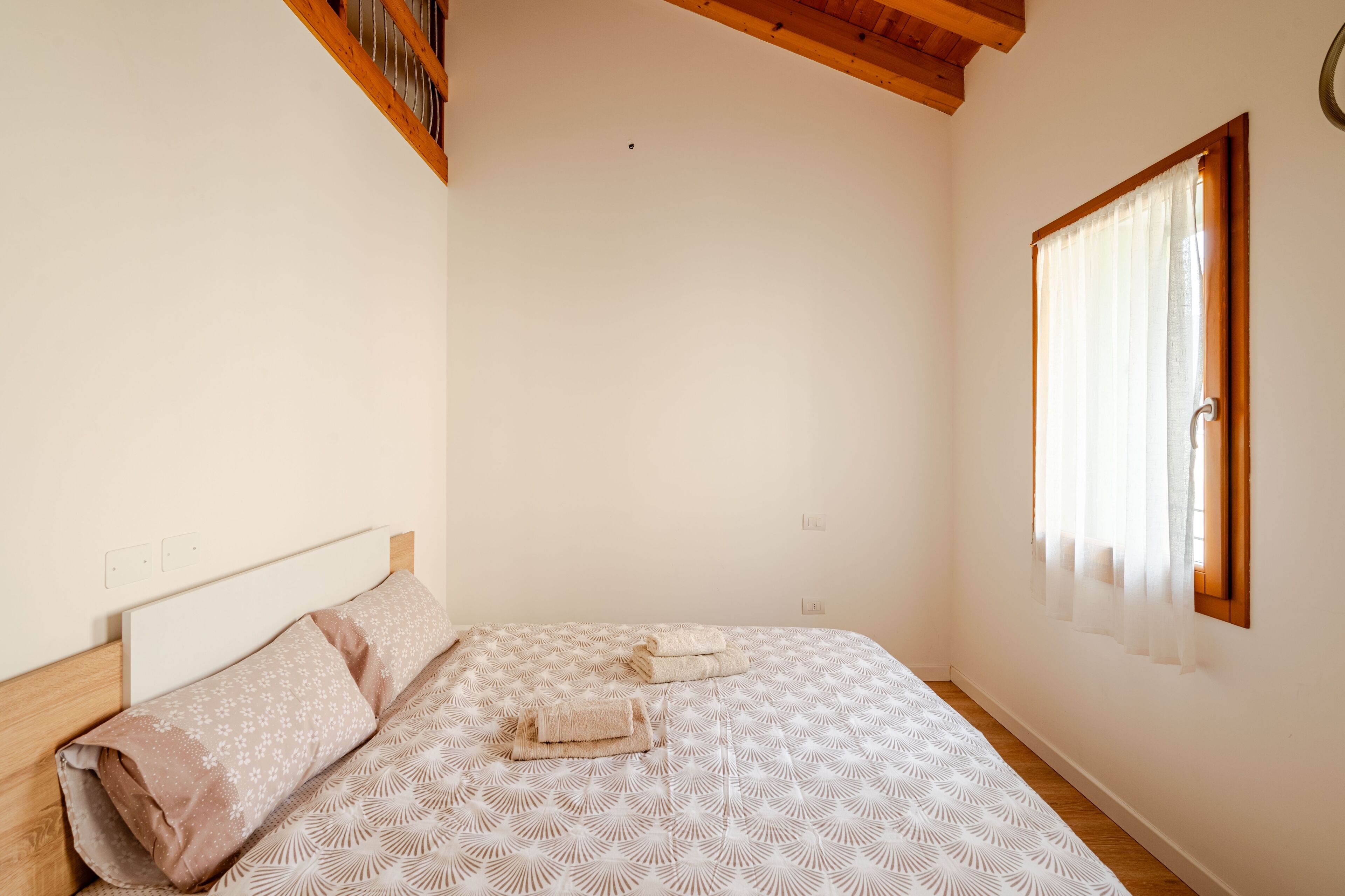 A bedroom with a bed, a wooden headboard, a window with curtains, and a ceiling with exposed beams.