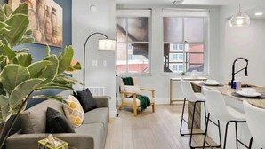Ett modernt vardagsrum med en soffa, en matplats med bord och stolar samt ett stort fönster med utsikt över byggnader.