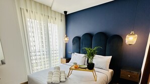 A modern bedroom with a large bed, wooden flooring, a dark blue accent wall, and a bedside table with a lamp. - Stay'Inn - Stadium Penthouse (Casablanca)