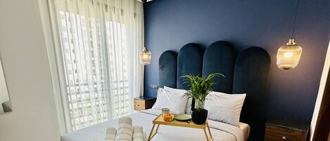 A modern bedroom with a large bed, wooden flooring, a dark blue accent wall, and a bedside table with a lamp.