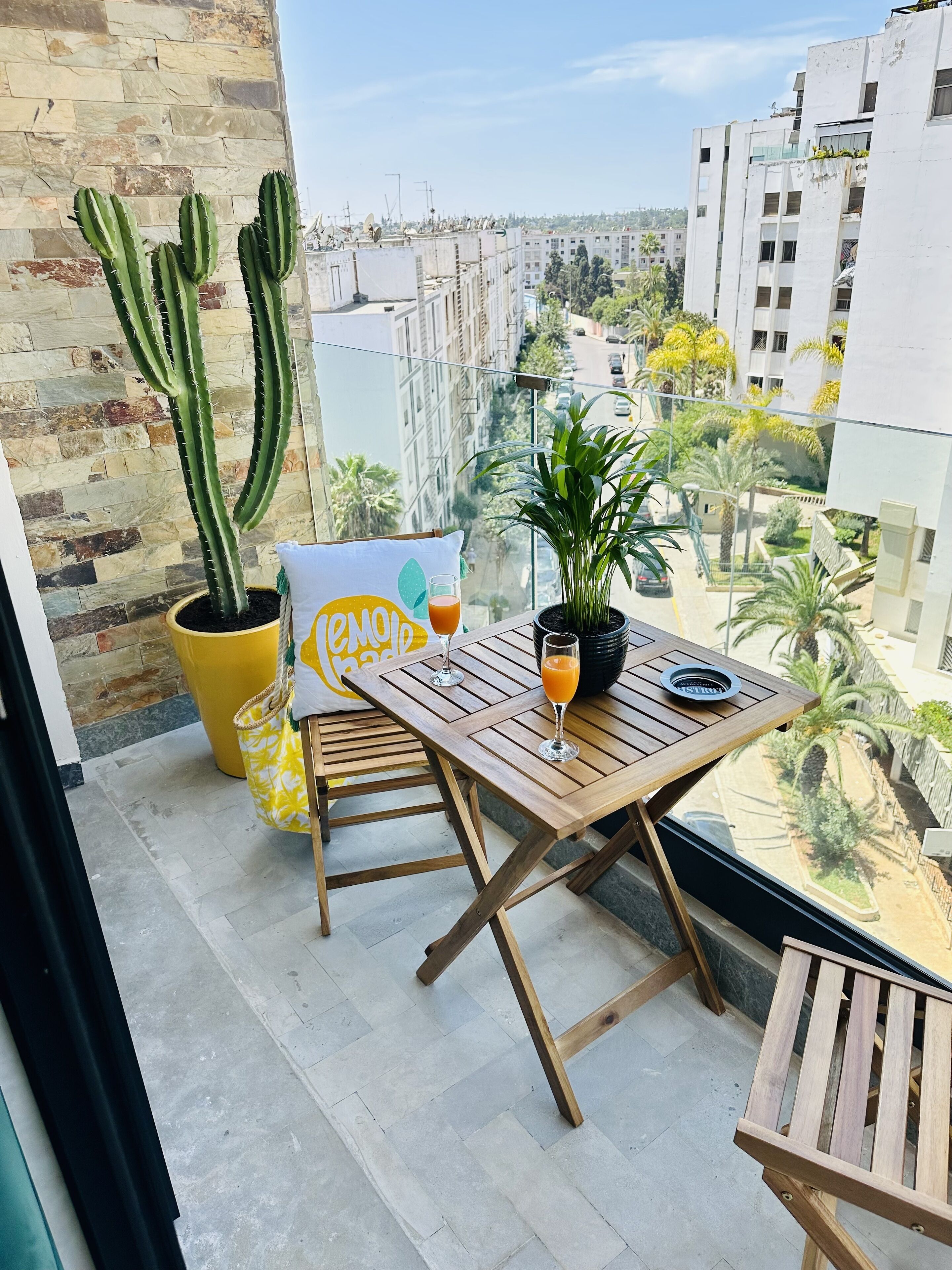 Een balkon met een houten tafel, twee stoelen, een cactus in pot, een bloempot en een glas sinaasappelsap.