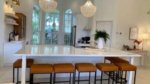 Una cocina amplia con una isla grande, taburetes de bar, luces colgantes y vistas amplias a través de grandes ventanales.
