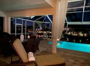 A poolside area with a view of the city at night, outdoor seating, and a covered patio. - Stunning SW home w/perfect indoor-outdoor living! Infinity pool/spa/great canal. (Cape Coral)