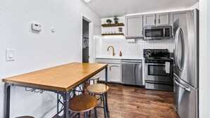 A compact kitchen with a wooden table, bar stools, stainless steel appliances, and white tiled backsplash.