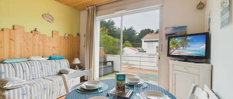 Una sala de estar acogedora con techo de madera, un sofá a rayas, una mesa de comedor preparada para cuatro personas y un televisor sobre un mueble.