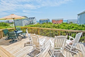 A wooden deck with outdoor furniture, including chairs and a table, overlooking a residential area with colorful houses and a view of the ocean. - Just Enough, Almost Oceanfront House in Emerald Isle, NC! (Emerald Isle)