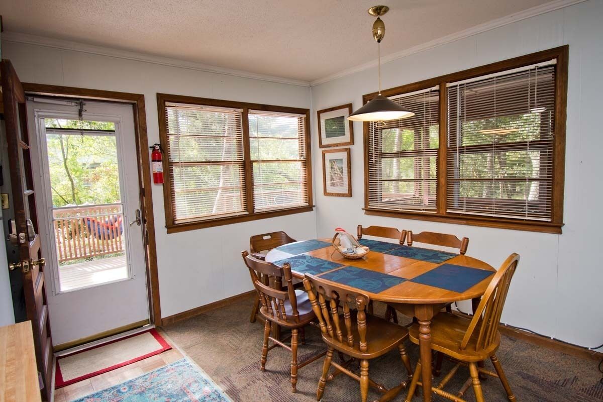 A dining area with a wooden table and chairs, large windows with blinds, a fire extinguisher, and a door with a red mat.