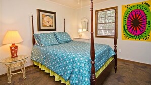 A bedroom with a four-poster bed, a ceiling fan, a bedside table with a lamp, a framed picture on the wall, and a large abstract painting. - Hasell Cottage, Oceanside House in Emerald Isle, NC! (Emerald Isle)