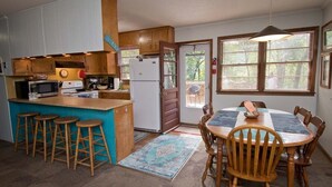 A compact kitchen with a central island, bar stools, a dining table with chairs, and a large window with blinds. - Hasell Cottage, Oceanside House in Emerald Isle, NC! (Emerald Isle)