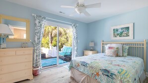 Une chambre avec un lit, une commode, une table de chevet, un ventilateur de plafond et une vue sur une piscine et un espace de détente extérieur.