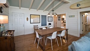 Un comedor con mesa de madera y sillas blancas, una lámpara colgante y una tabla de surf en la pared.