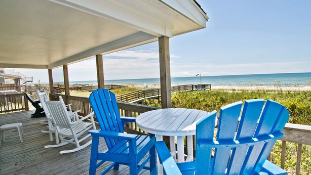 Una terraza de madera con sillas Adirondack azules y una mesa blanca, con vistas a una playa que tiene un paseo de madera y un muelle.