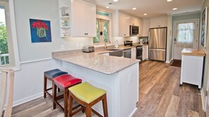 A modern kitchen with white cabinetry, granite countertops, and stainless steel appliances. There are three stools with colorful cushions. - Hollandaise, Oceanview House in Emerald Isle, NC! (Emerald Isle)