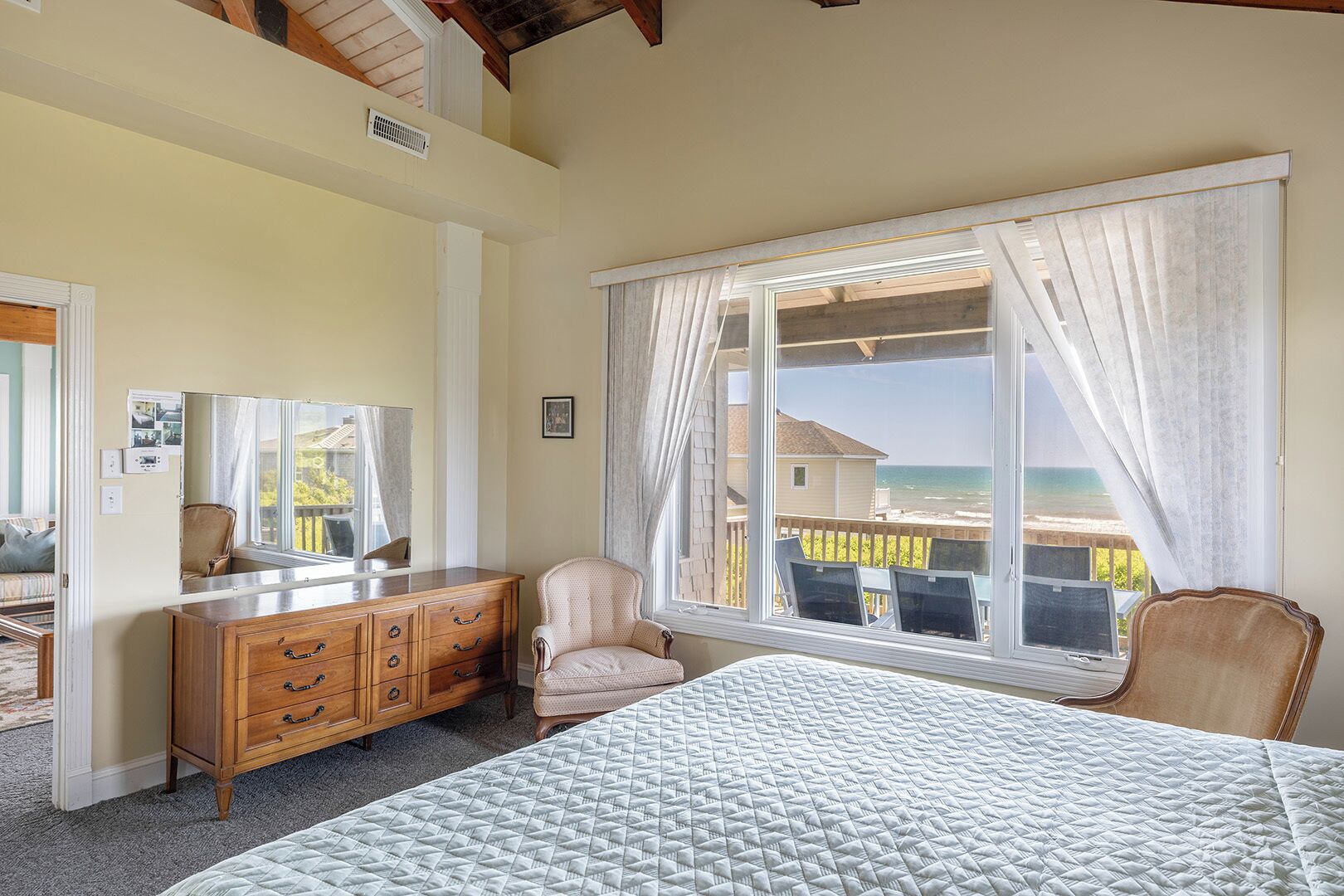 A bedroom with a bed, a dresser, a chair, and a large window with a view of the ocean.