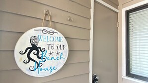 A door with a sign that reads "WELCOME TO OUR Beach House" on the wall. - Island Beach Racquet Club 105B, Oceanside Condo in Atlantic Beach, NC! (Atlantic Beach)