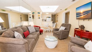 A living room with a sofa set, a glass coffee table, a dining area with white chairs, and a TV displaying a beach scene. - Island Beach Racquet Club 303A, Oceanfront Condo in Atlantic Beach, NC! (Atlantic Beach)
