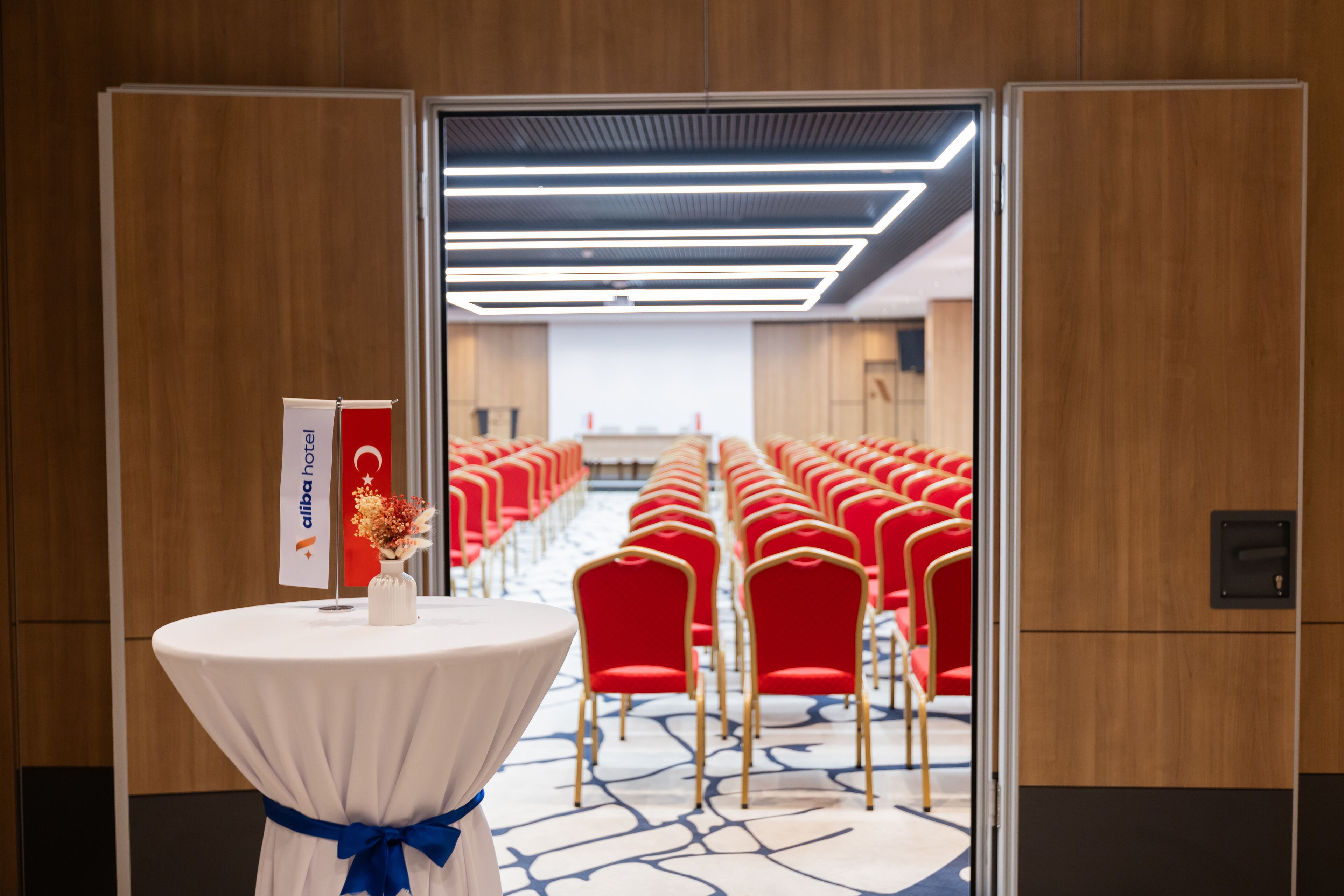 A hotel room with a view of a conference room with red chairs and a white table with a blue ribbon.