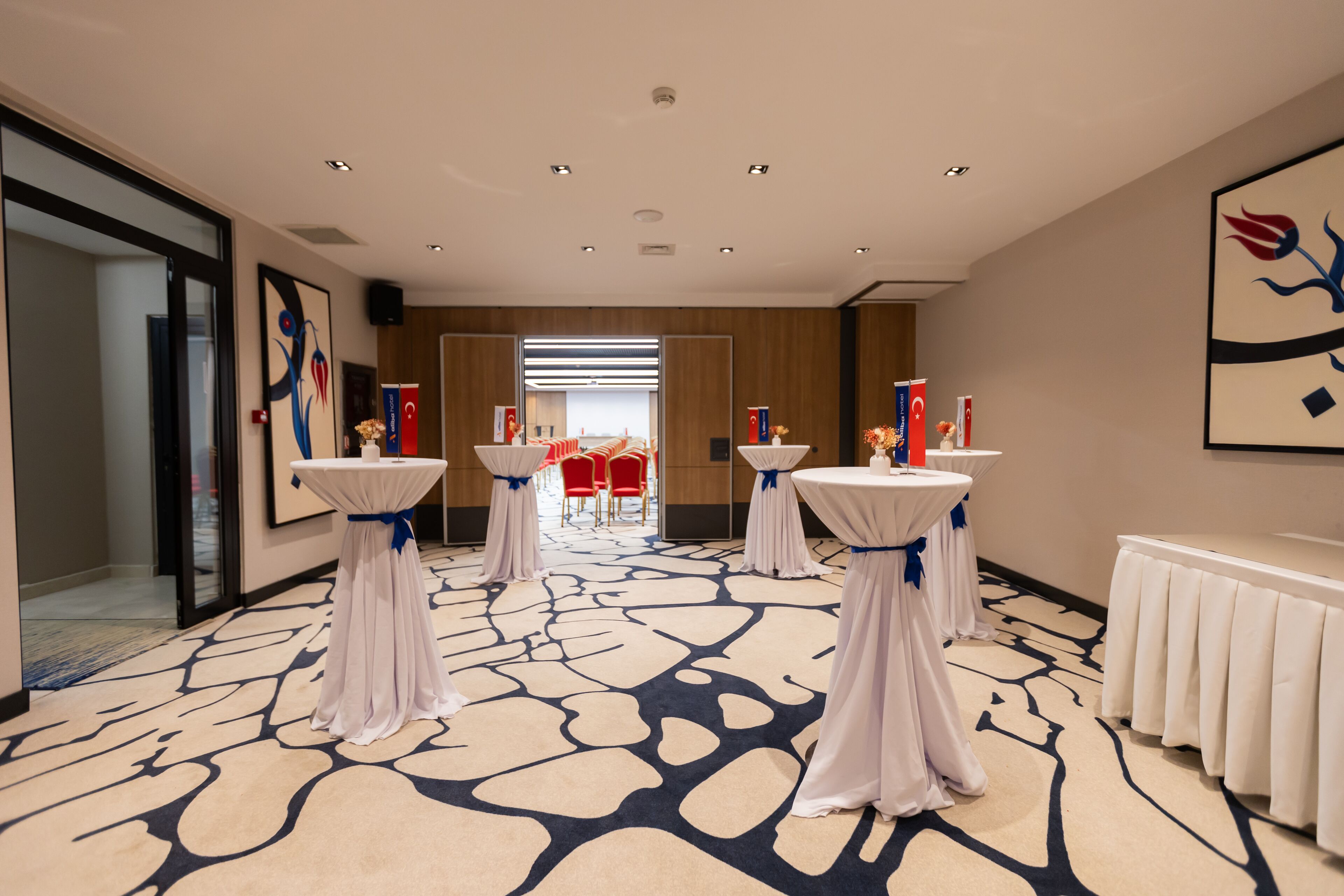 A hotel lobby with a modern design, featuring a patterned carpet, white tables with blue sashes, and red chairs in the background.
