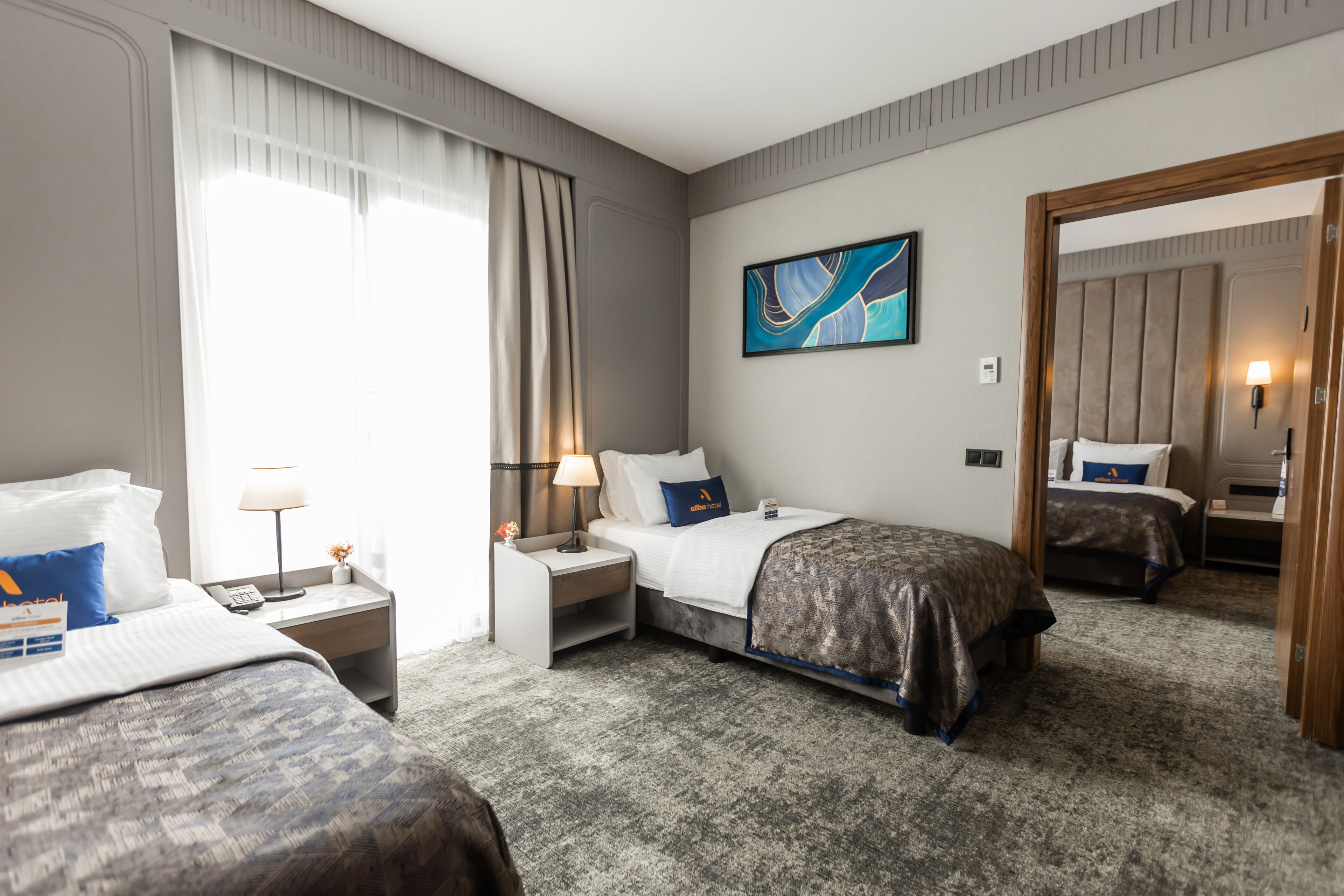 Habitación de hotel con dos camas, una mesita de noche con lámpara, un cuadro en la pared y piso alfombrado.