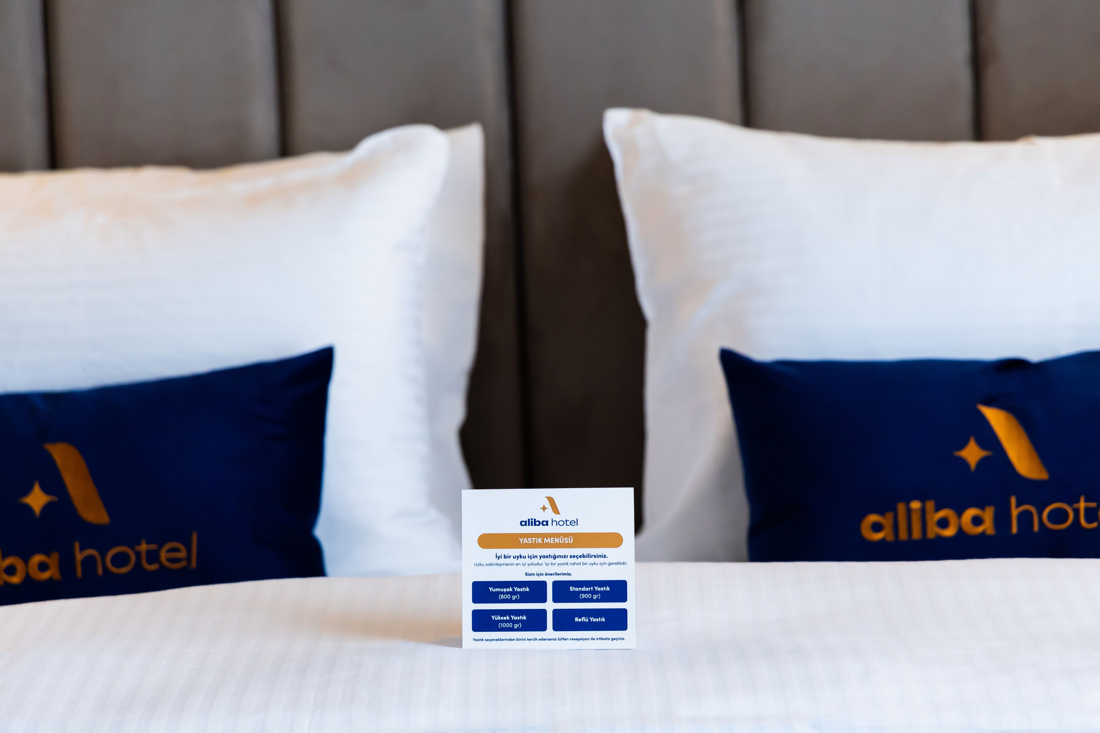 A hotel bed with pillows labeled "Alibaba Hotel" and a card detailing room amenities.