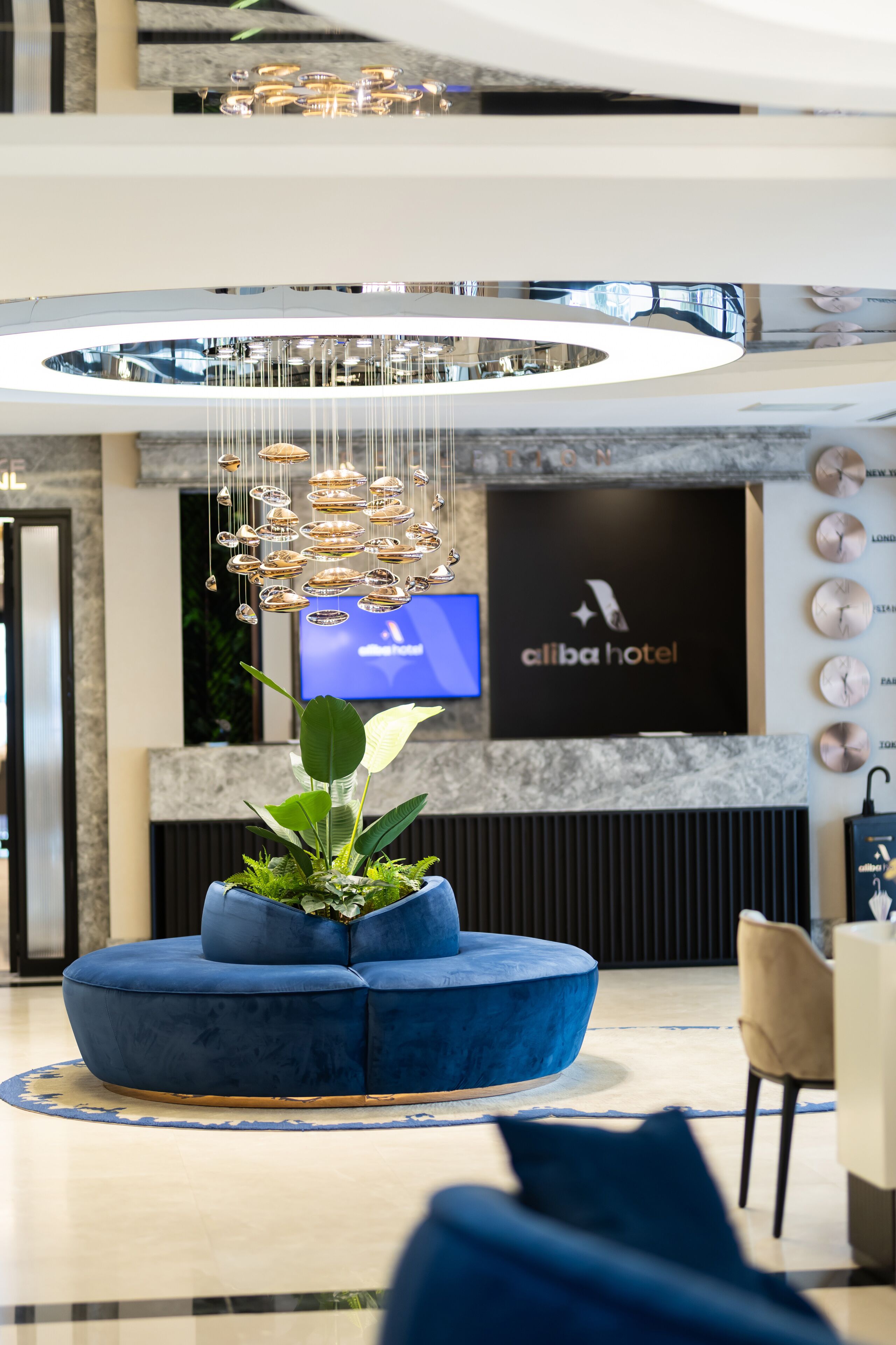 Een moderne hotel lobby met een blauwe zithoek, een grote kroonluchter en een televisiescherm waarop de naam van het hotel staat.