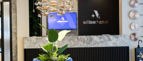 Een moderne hotel lobby met een blauwe zithoek, een grote kroonluchter en een televisiescherm waarop de naam van het hotel staat.