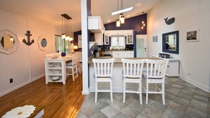 A modern kitchen with white cabinets, a dining area with white chairs, and a large island. - Knot A Care, Oceanside House in Emerald Isle, NC! (Emerald Isle)