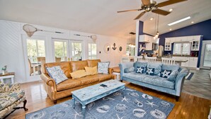 A spacious living room with a brown leather sofa, a blue sofa, and a wooden coffee table. There is a ceiling fan, a kitchen area with white cabinets, and a large window. - Knot A Care, Oceanside House in Emerald Isle, NC! (Emerald Isle)