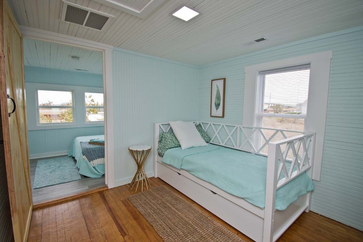 A bedroom with a white bed, a small round table, a window with blinds, and a view of the outdoors.