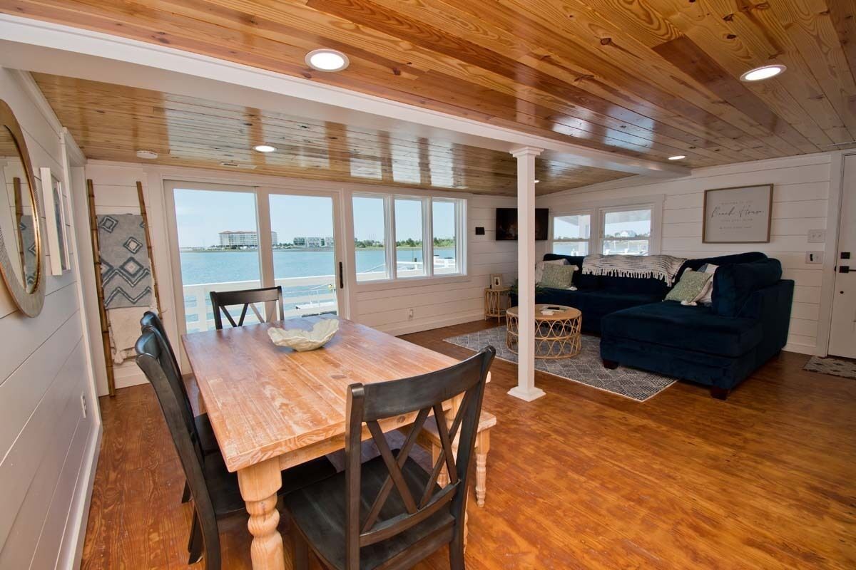 A spacious living area with a wooden dining table, dark blue sofa, and large windows overlooking a body of water.