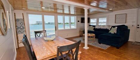A spacious living area with a wooden dining table, dark blue sofa, and large windows overlooking a body of water.