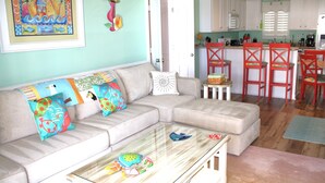 A living room with a beige sectional sofa, colorful pillows, a glass coffee table with toy fish, and a kitchen area with white cabinets and red chairs. - License to Chill, Almost Oceanfront House in Emerald Isle, NC! (Emerald Isle)