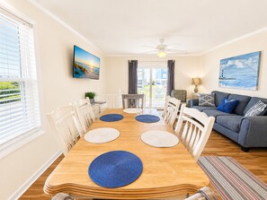 A dining area with a wooden table, white chairs, and blue placemats. - Living Under The Rainbow, Almost Oceanfront House in Emerald Isle, NC! (Emerald Isle)