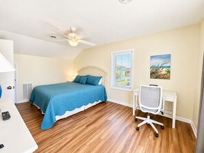 A bedroom with a bed, a desk, a chair, a ceiling fan, a window with blinds, and a painting on the wall. - Living Under The Rainbow, Almost Oceanfront House in Emerald Isle, NC! (Emerald Isle)