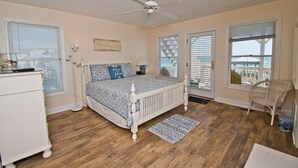 Ein Schlafzimmer mit einem Bett, einem Deckenventilator, einem Fenster mit Jalousien, einem Sessel und Blick ins Freie.
